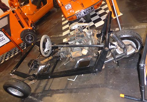 Black ladder chassis with an engine, steering wheel and three small wheels on a show floor. Opens a larger view