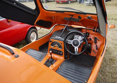 Orange cabin with a black instrument binnacle, small steering wheel and rubber floor mats. Opens a larger view