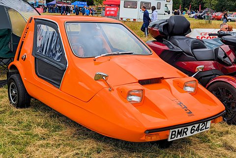 Tangerine three-wheeler on dry grass beside a red touring motorcycle, show tents behind. Opens a larger view