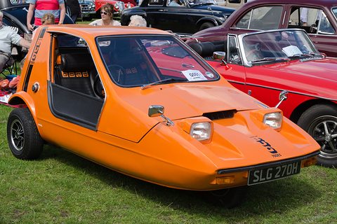 Tangerine three-wheeler with side screens off, parked on grass beside a red sports car.