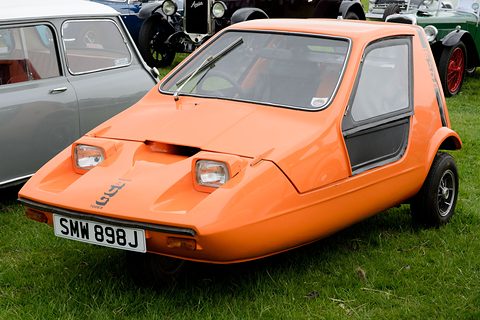 Tangerine three-wheeler on grass between a grey Mini and other classic cars. Opens a larger view