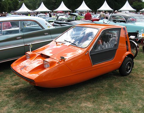 Tangerine three-wheeler on a show lawn among classic cars, white marquees behind. Opens a larger view