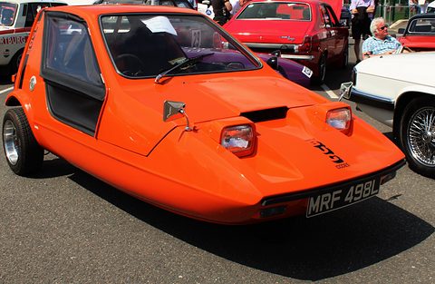 Tangerine three-wheeler on tarmac in bright sun, classic cars and spectators behind. Opens a larger view