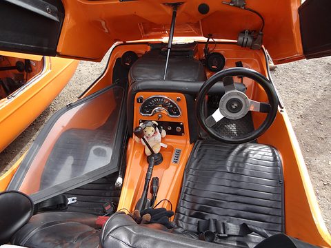 Orange cabin with black ribbed seats, a three-spoke wheel and a small doll on the centre console. Opens a larger view