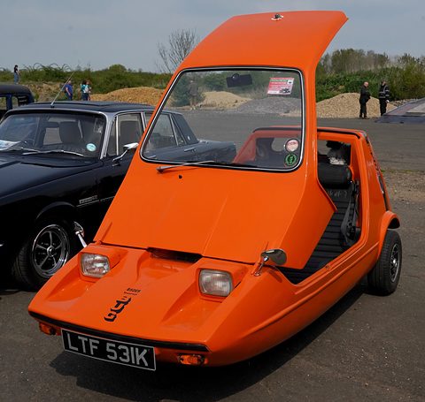 Tangerine three-wheeler head-on with its canopy lifted, beside a dark saloon under a grey sky. Opens a larger view