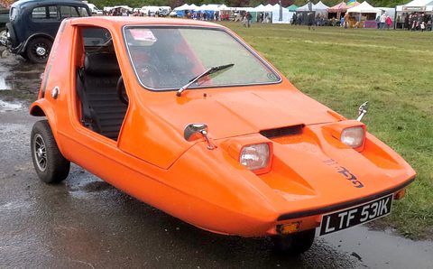 Tangerine three-wheeler on a wet hardstanding, stalls and caravans behind. Opens a larger view
