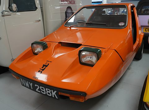 Tangerine three-wheeler on a grey museum floor, a cream van beside it. Opens a larger view