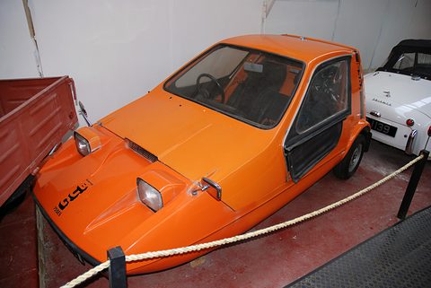 Tangerine three-wheeler on a dark floor behind a rope, a white sports car alongside. Opens a larger view
