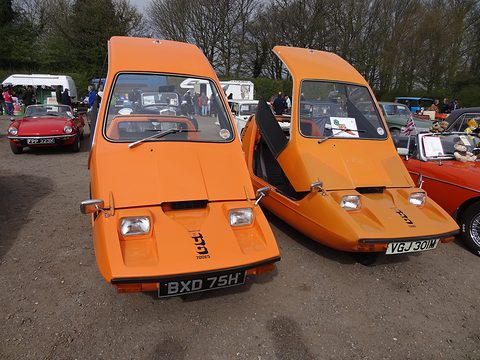 Two tangerine three-wheelers facing the camera on gravel, bare trees and other cars behind. Opens a larger view