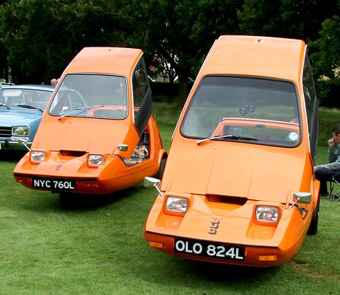 Two tangerine wedge three-wheelers side by side facing the camera on a grass field, trees behind. Opens a larger view