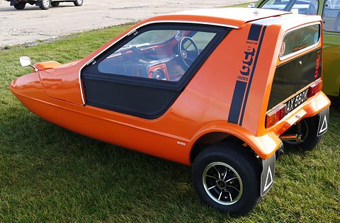 Tangerine three-wheeler from the rear quarter on grass, black stripe and warning triangle on the mud flap. Opens a larger view