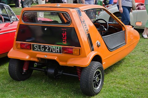 Tangerine three-wheeler from the rear quarter on grass, red rear dampers visible under the tail. Opens a larger view