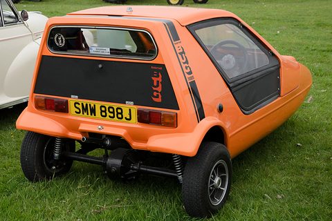 Tangerine three-wheeler from behind with a yellow plate, black tail panel and coil springs visible. Opens a larger view