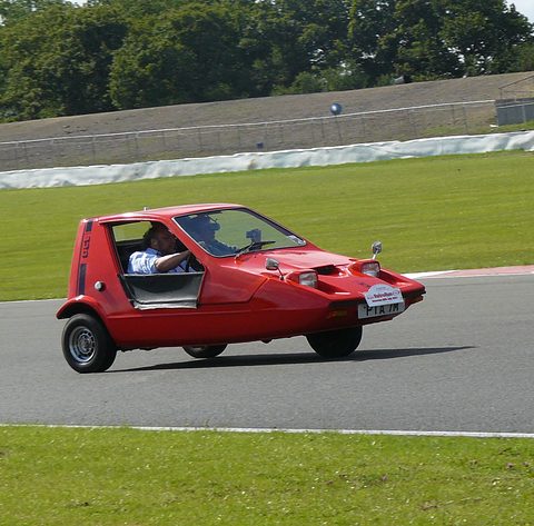 Red wedge three-wheeler with its driver visible, cornering on a track with grass and stands behind. Opens a larger view