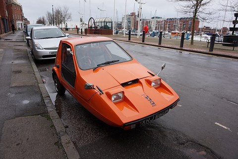 Tangerine three-wheeler at a kerb on a rain-wet street beside a marina. Opens a larger view
