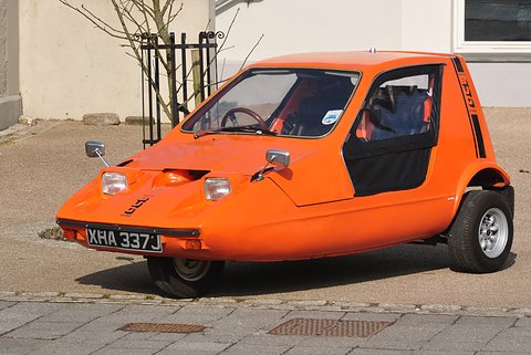 Tangerine three-wheeler parked on a paved forecourt in front of a white building. Opens a larger view
