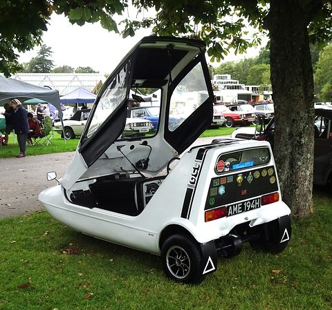 White three-wheeler from the rear quarter on grass, stickers in its rear window and the canopy lifted. Opens a larger view