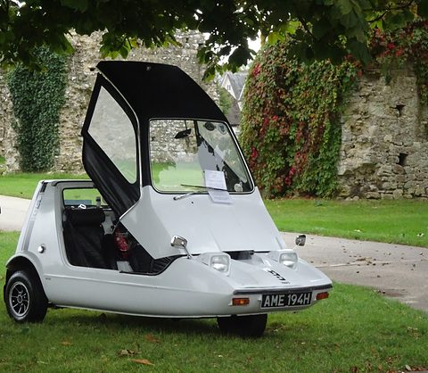 White wedge three-wheeler with its canopy open, parked on grass before an ivy-covered stone wall.