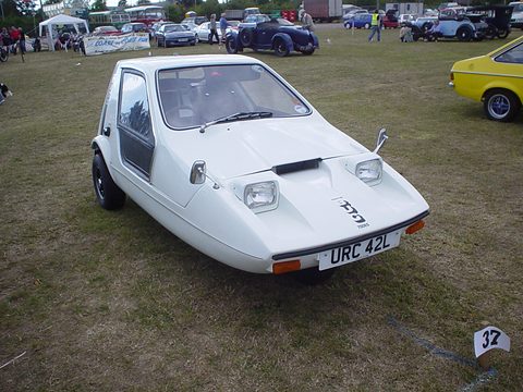 White wedge three-wheeler parked on a grass show field, buses and a banner behind. Opens a larger view
