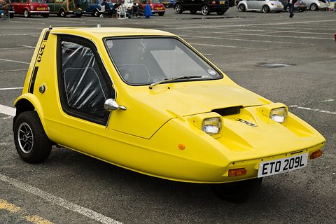 Yellow wedge three-wheeler on a car park, black side screens fitted. Opens a larger view