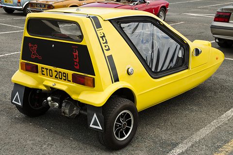 Yellow three-wheeler from behind with a black tail panel, stripes and warning triangles on the mud flaps. Opens a larger view