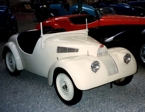 Cream rounded roadster with a horseshoe grille on grey gravel, a red racing car behind. Opens a larger view