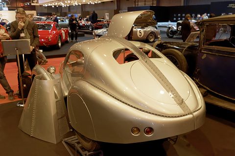 Rear of a silver teardrop coupé with a central spine and a raised gullwing door. Opens a larger view