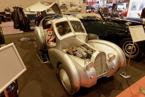 Silver streamlined miniature coupé with its bonnet open and gullwing doors raised on a show stand.