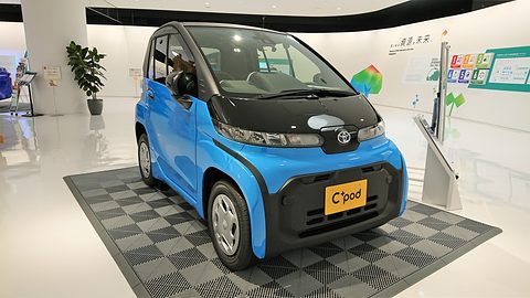 Bright blue tiny two-seat car with a black roof and front, on a grey chequered display mat in a white showroom. Opens a larger view