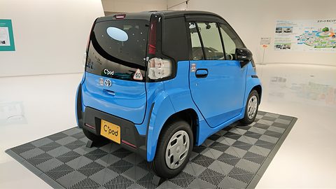 Rear of a bright blue tiny two-seat car with a black tailgate on a chequered mat in a white showroom. Opens a larger view