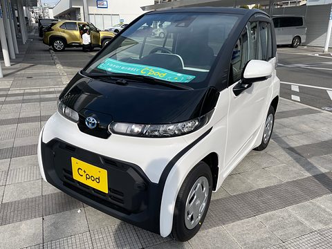 White tiny two-seat car with a black roof and a C+pod plate on dealer paving, other cars behind.