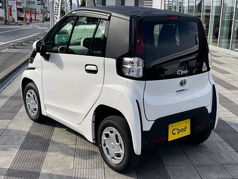 Rear of a white tiny two-seat car with a black tailgate and a C+pod plate, on grey paving. Opens a larger view