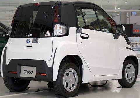 Rear of a white tiny two-seat car with a black tailgate and a C+pod plate, on charge in a showroom. Opens a larger view