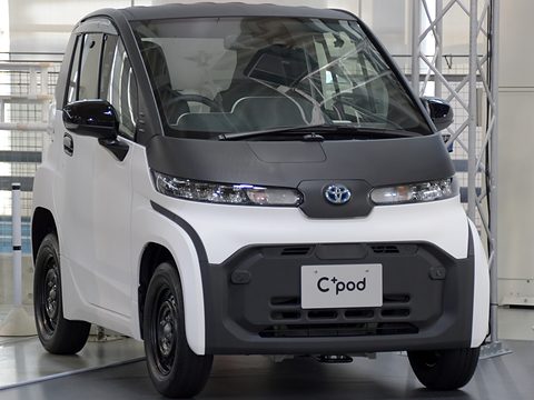 Front of a white tiny two-seat car with black steel wheels and a C+pod plate under a metal display frame.