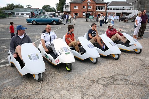 Five people in a row of white electric tricycles on a tarmac yard. Opens a larger view