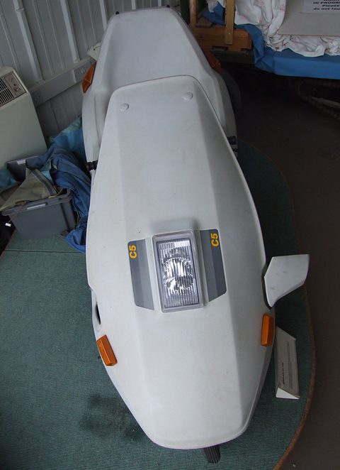 White electric tricycle facing the camera on a green carpeted plinth. Opens a larger view