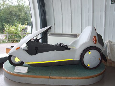White electric tricycle side-on on a round green plinth inside a corrugated shed. Opens a larger view