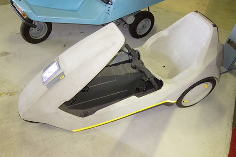 White electric tricycle seen from above on a pale floor, a blue vehicle behind. Opens a larger view
