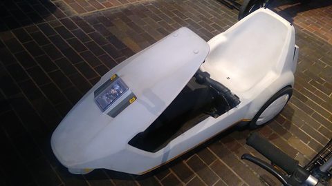 White electric tricycle on a brick floor in dim gallery light. Opens a larger view