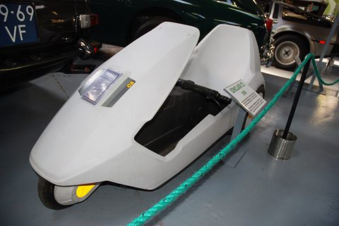 White electric tricycle on a grey floor behind a green rope, classic cars behind. Opens a larger view