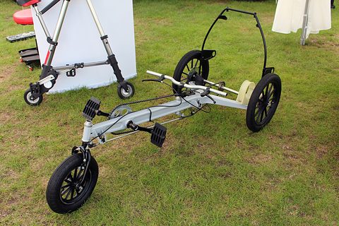 Silver tubular frame with pedals, a handlebar and three spoked wheels standing on grass. Opens a larger view