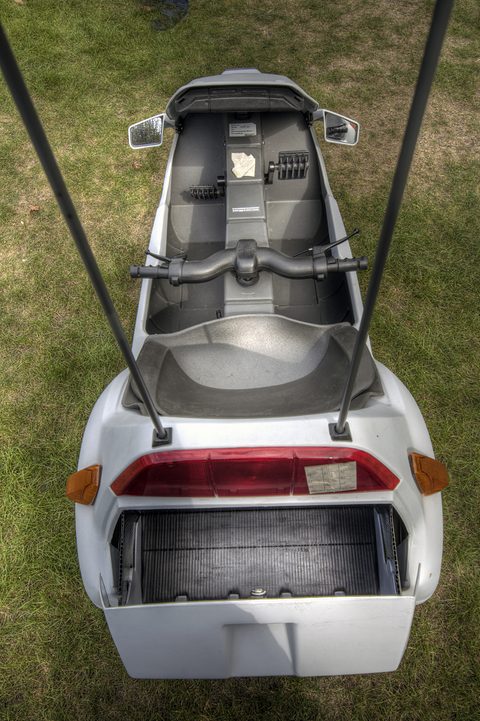 Looking down on an open electric tricycle: seat, handlebar and an empty rear compartment, on grass. Opens a larger view