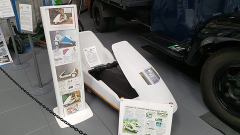 White electric tricycle beside a display stand of brochures and leaflets, behind a chain. Opens a larger view