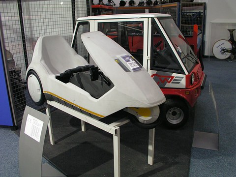 White electric tricycle raised on a frame in front of a small white and red car. Opens a larger view