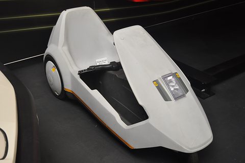 White electric tricycle with its nose lamp, on a dark display floor. Opens a larger view