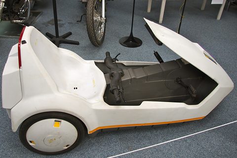 White electric tricycle side-on on blue carpet, its nose lifted open, a motorcycle wheel behind. Opens a larger view