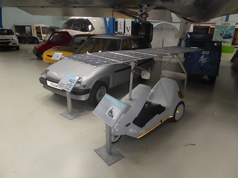 White electric tricycle under a raised frame of solar panels, small cars displayed behind. Opens a larger view