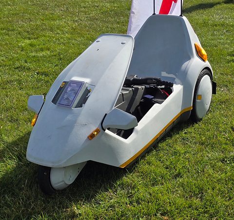 White single-seat electric tricycle on grass, its open cockpit and black seat visible. Opens a larger view
