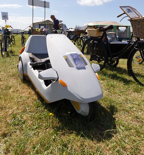 White electric tricycle on a show field beside a row of old bicycles. Opens a larger view