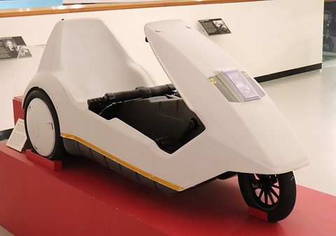 White electric tricycle on a red display plinth in a bright gallery. Opens a larger view